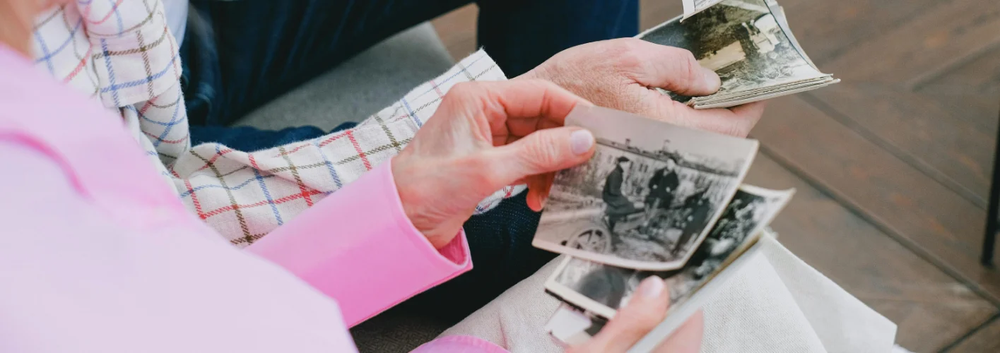 Two older adults sitting together, holding and looking through a collection of black and white photographs, sharing memories and providing dementia family support.